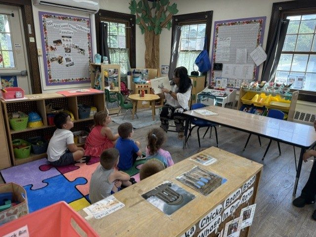 A teacher reading a picture book to children seated on a colorful puzzle rug