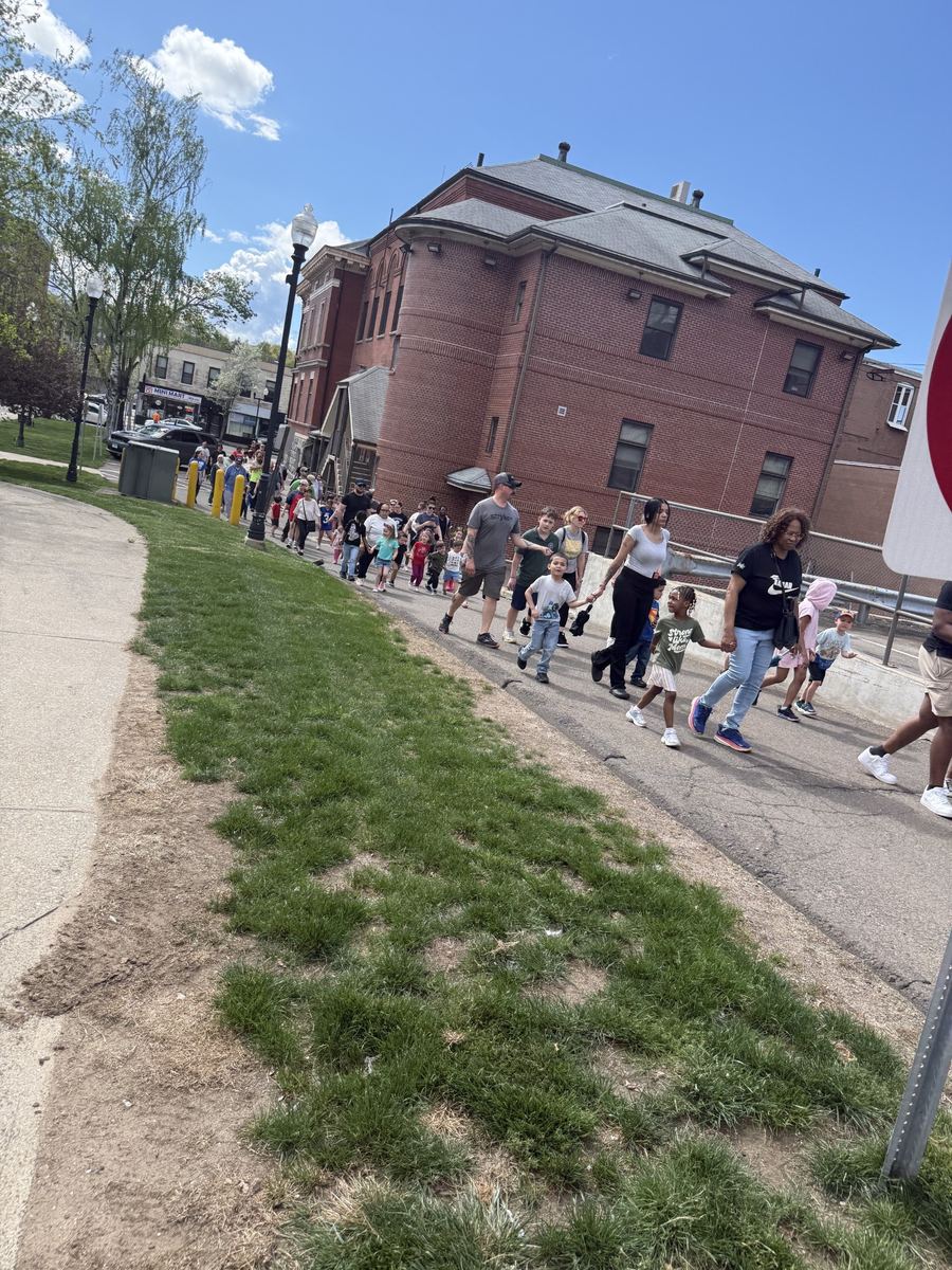 Children, families and staff walking together along a sidewalk past a red brick building on the Family Walk