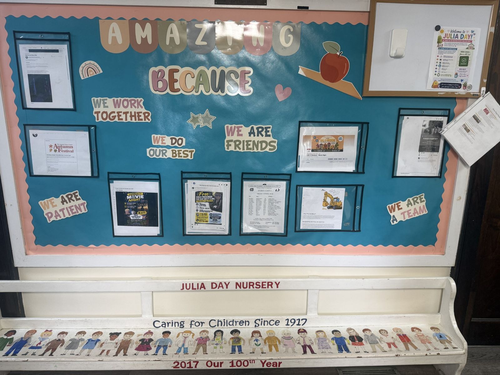 A white wooden bench painted with a row of children and the words Julia Day Nursery, Caring for Children Since 1917, beneath a classroom bulletin board headed Amazing Because