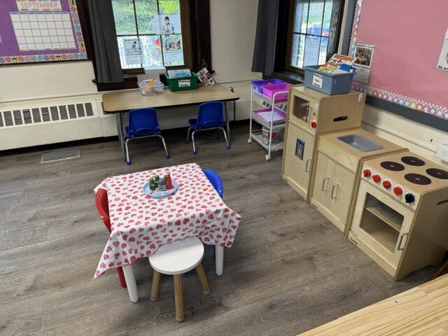 A classroom corner with a wooden play kitchen, a small table set with a strawberry-print cloth, and a September calendar on the wall