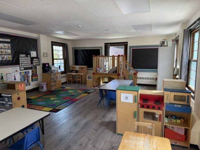 A wide view of a classroom with an alphabet rug, a wooden loft climber, a play kitchen and child-sized tables and chairs