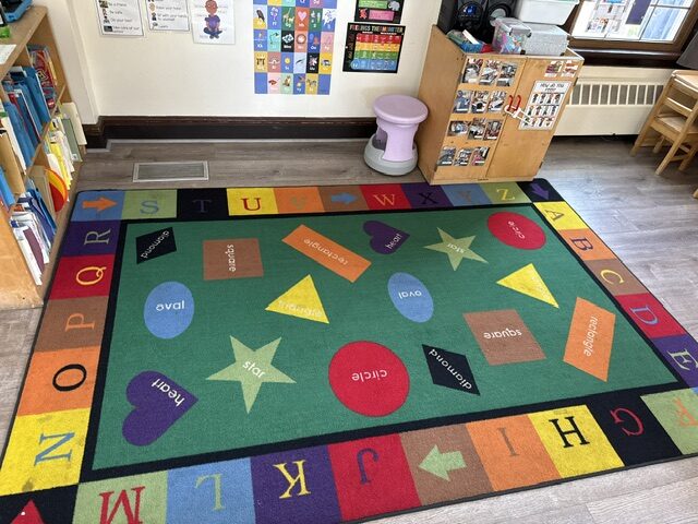 A colorful alphabet and shapes rug beside low bookshelves in a classroom
