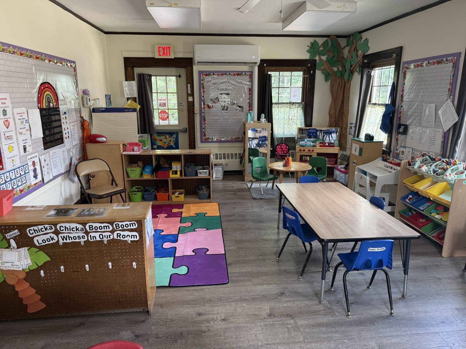 A wide classroom with a colorful puzzle rug, child-sized tables with blue chairs and a Chicka Chicka Boom Boom display beside the door