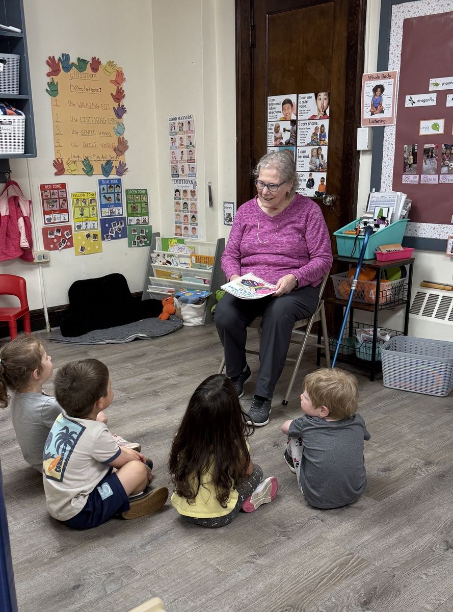 A visitor from the senior center reading a picture book to children seated on the classroom floor