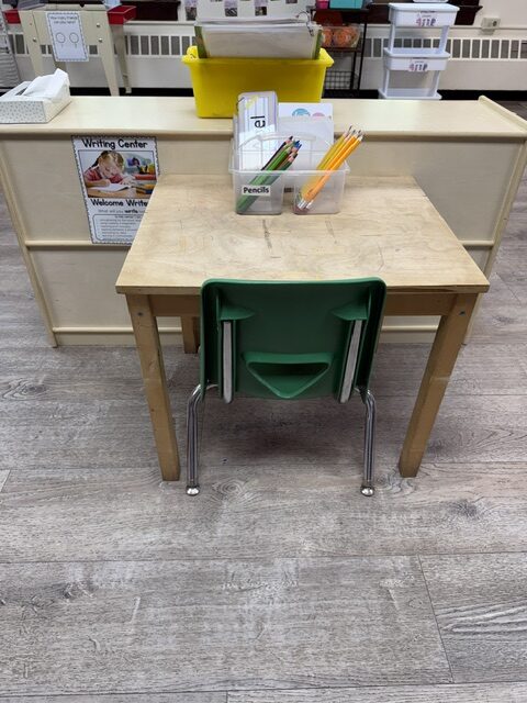 A small wooden table with a green chair and a jar of pencils at the Writing Center, under a sign reading Welcome Writers